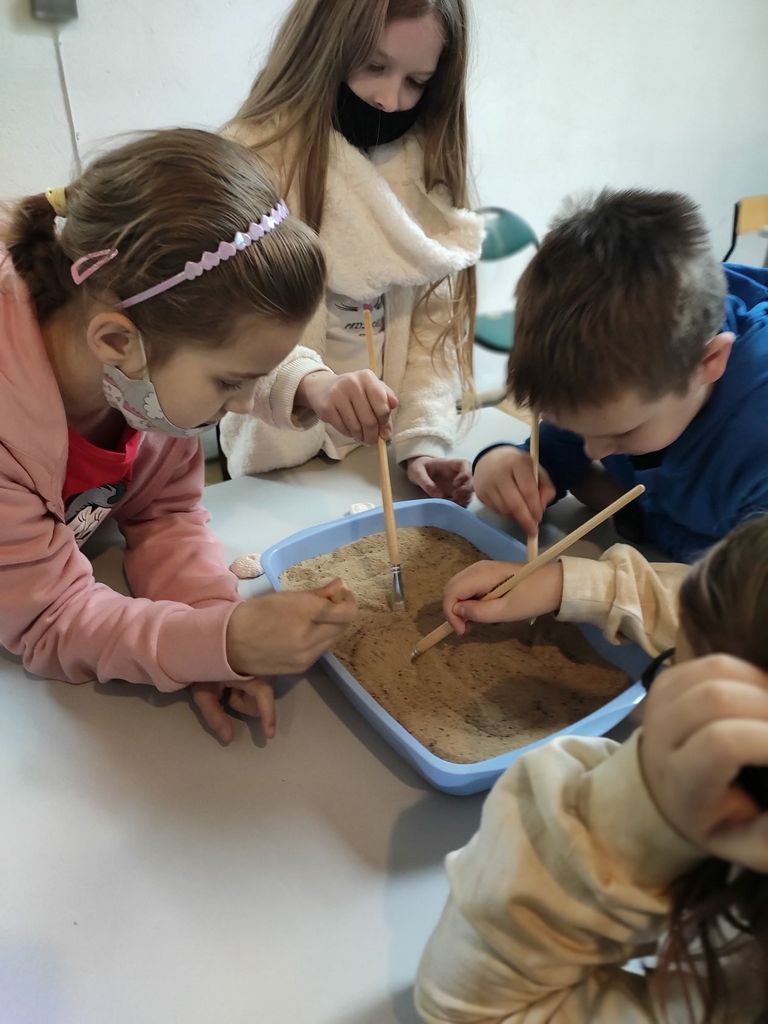 Dzieci przy kuwecie z piaskiem próbują wykopać skamielinę